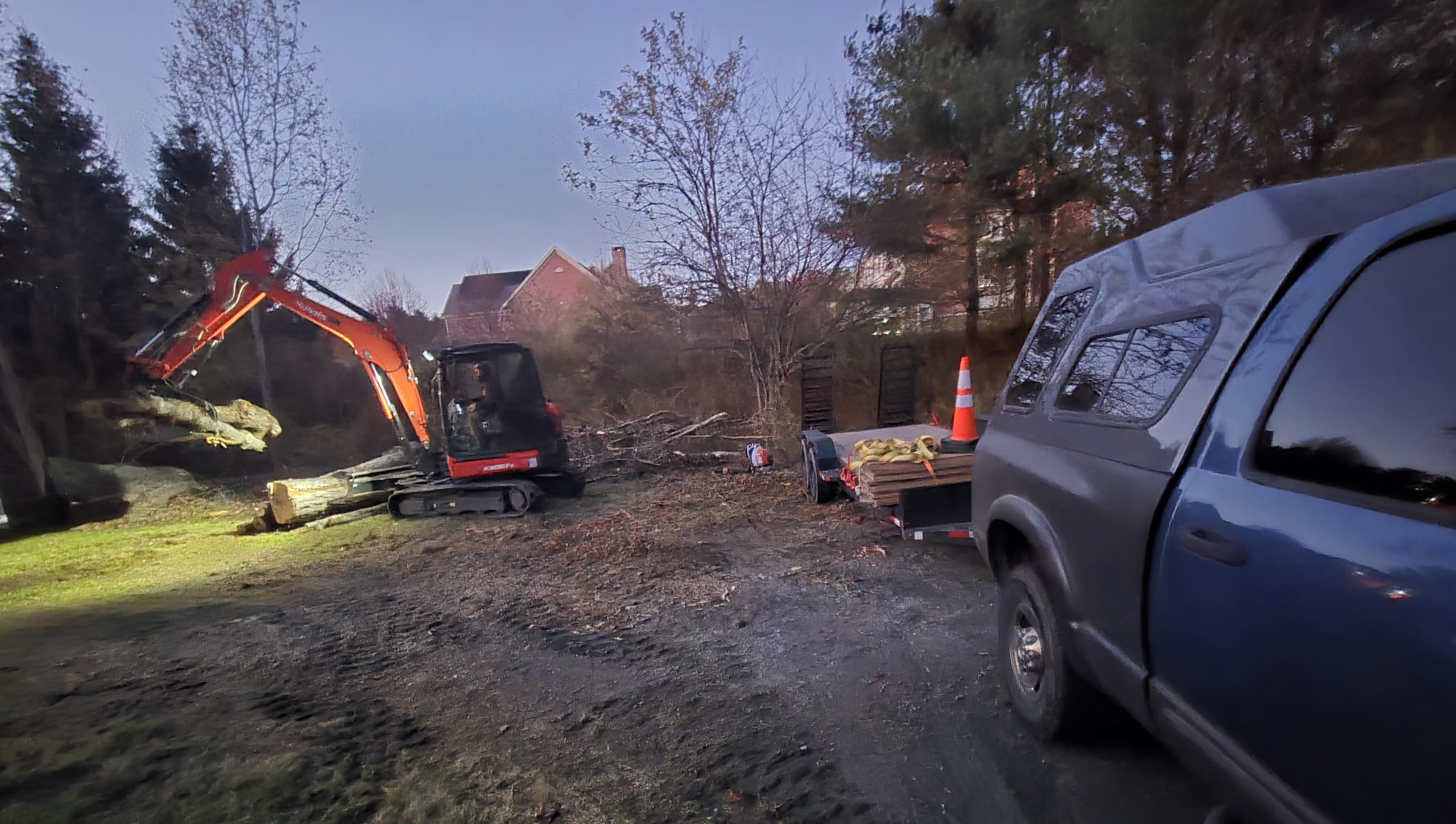 Evening excavation site with an orange compact excavator near logs and stumps, traffic cone and pickup truck nearby, golden-hour light and residential structures in the background.